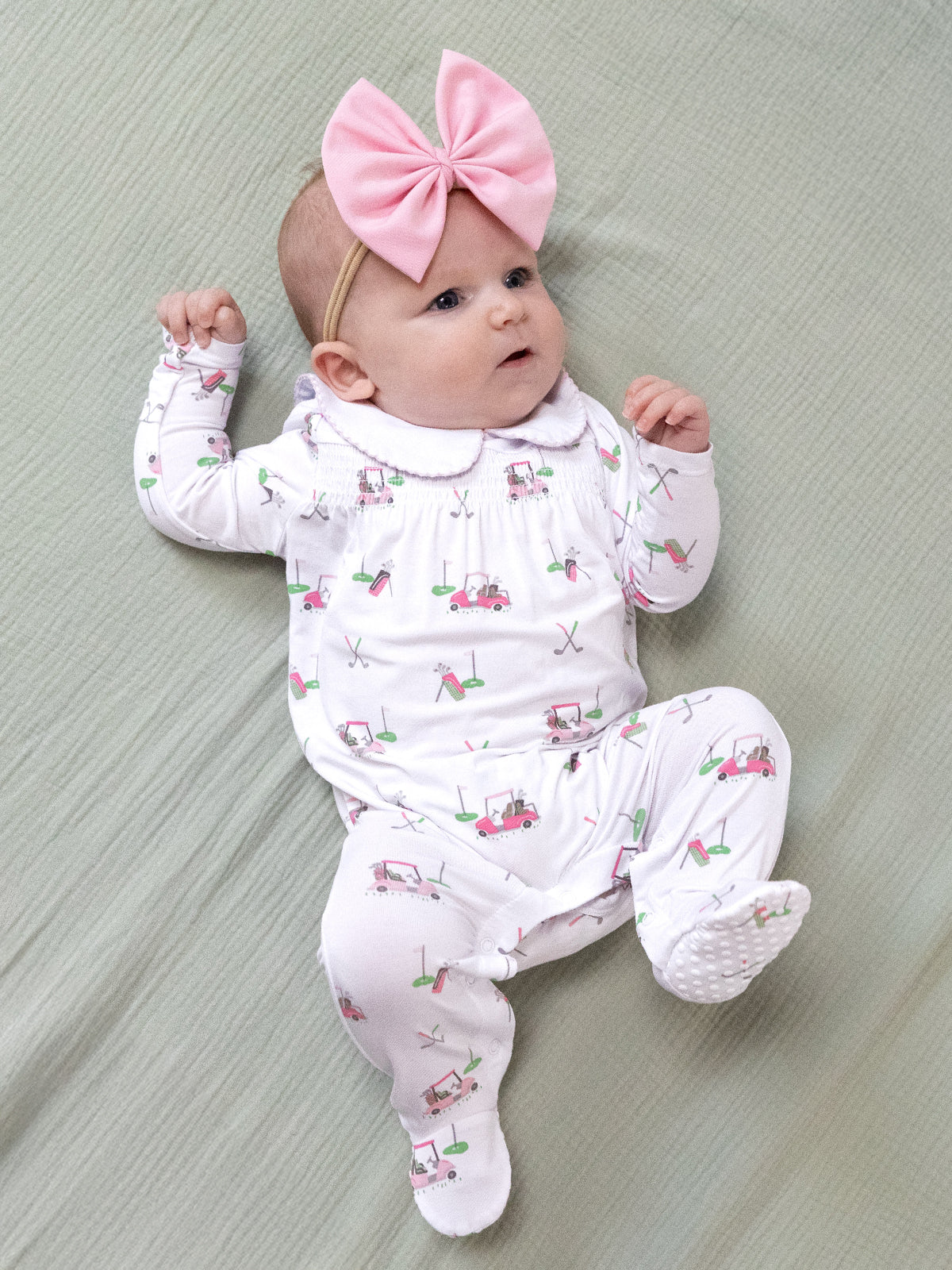 Infant girl in a golf-themed outfit and pink bow, lying on a soft green blanket, smiling curiously.
