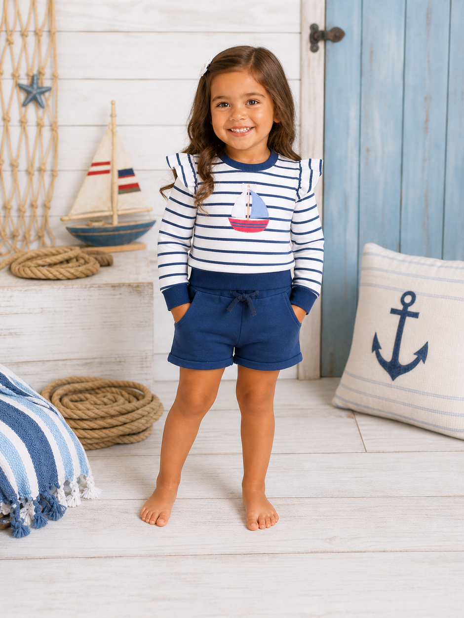 Smiling girl in a striped shirt with a sailboat design, wearing navy shorts, standing in a nautical-themed room.