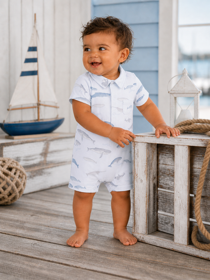Smiling toddler in whale-patterned romper, standing by wooden crate and sailboat decor on a sunny, outdoor setting.