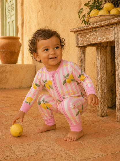 Smiling child in lemon-patterned pajamas squatting beside a lemon on a rustic outdoor surface.