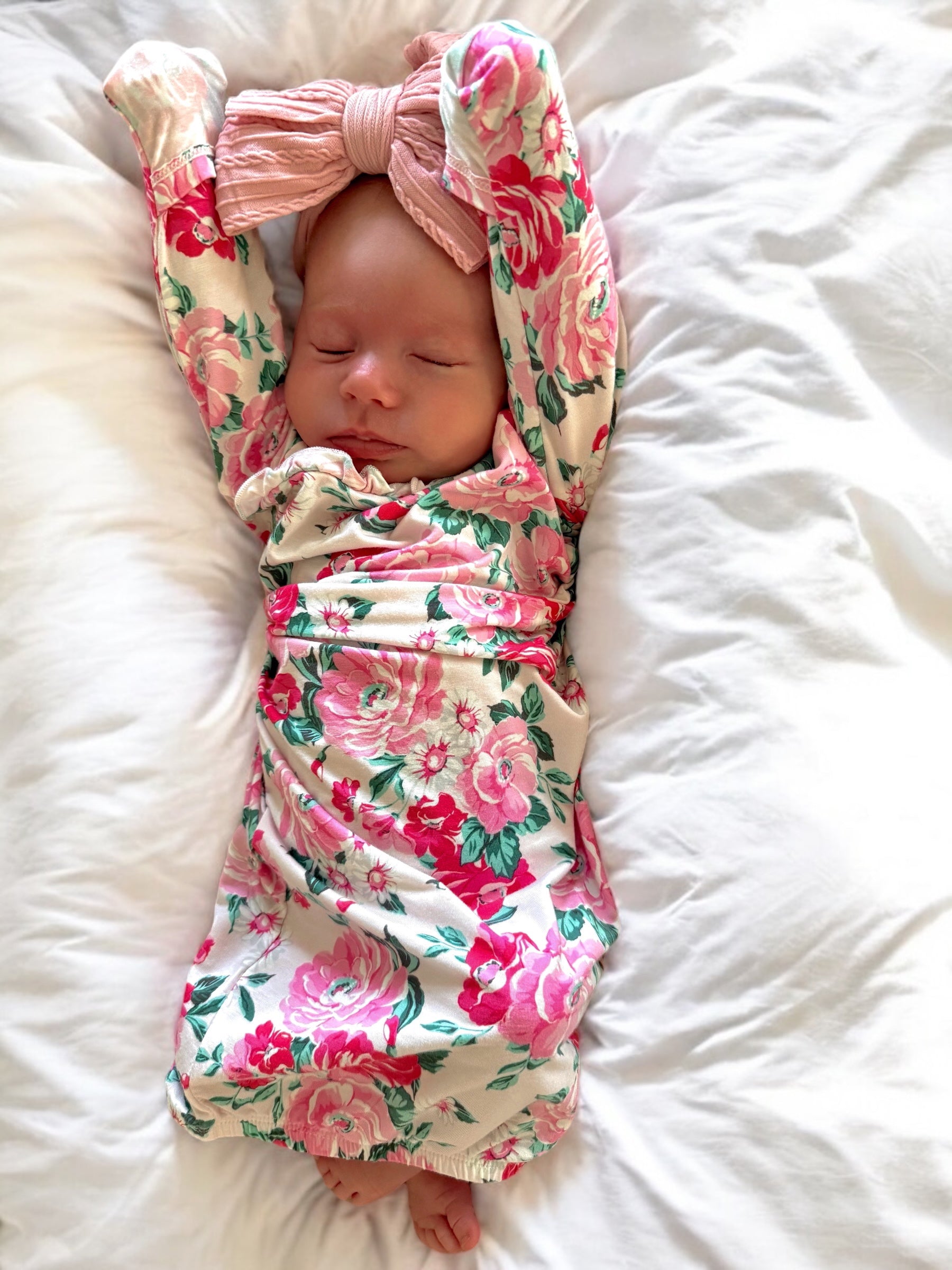 Newborn baby sleeping on white blankets, wearing a floral outfit and a large pink bow headband.