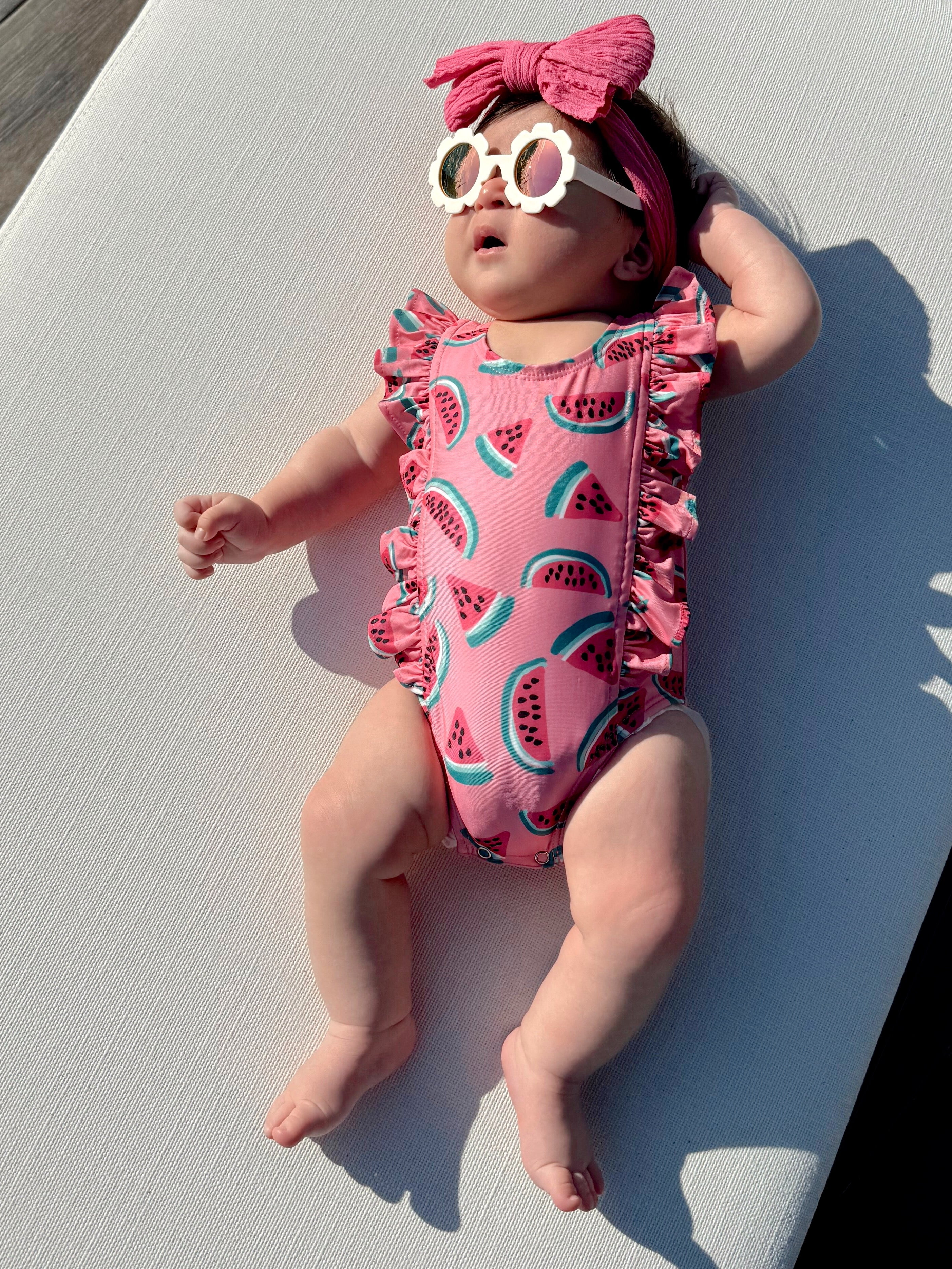 Baby wearing a pink watermelon-patterned swimsuit, sunglasses, and headband, lying on a sunlit surface.