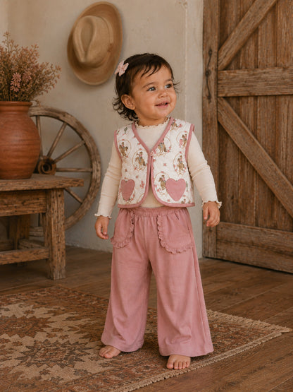 Smiling toddler in a floral vest and pink pants, standing on a rug in a rustic setting.