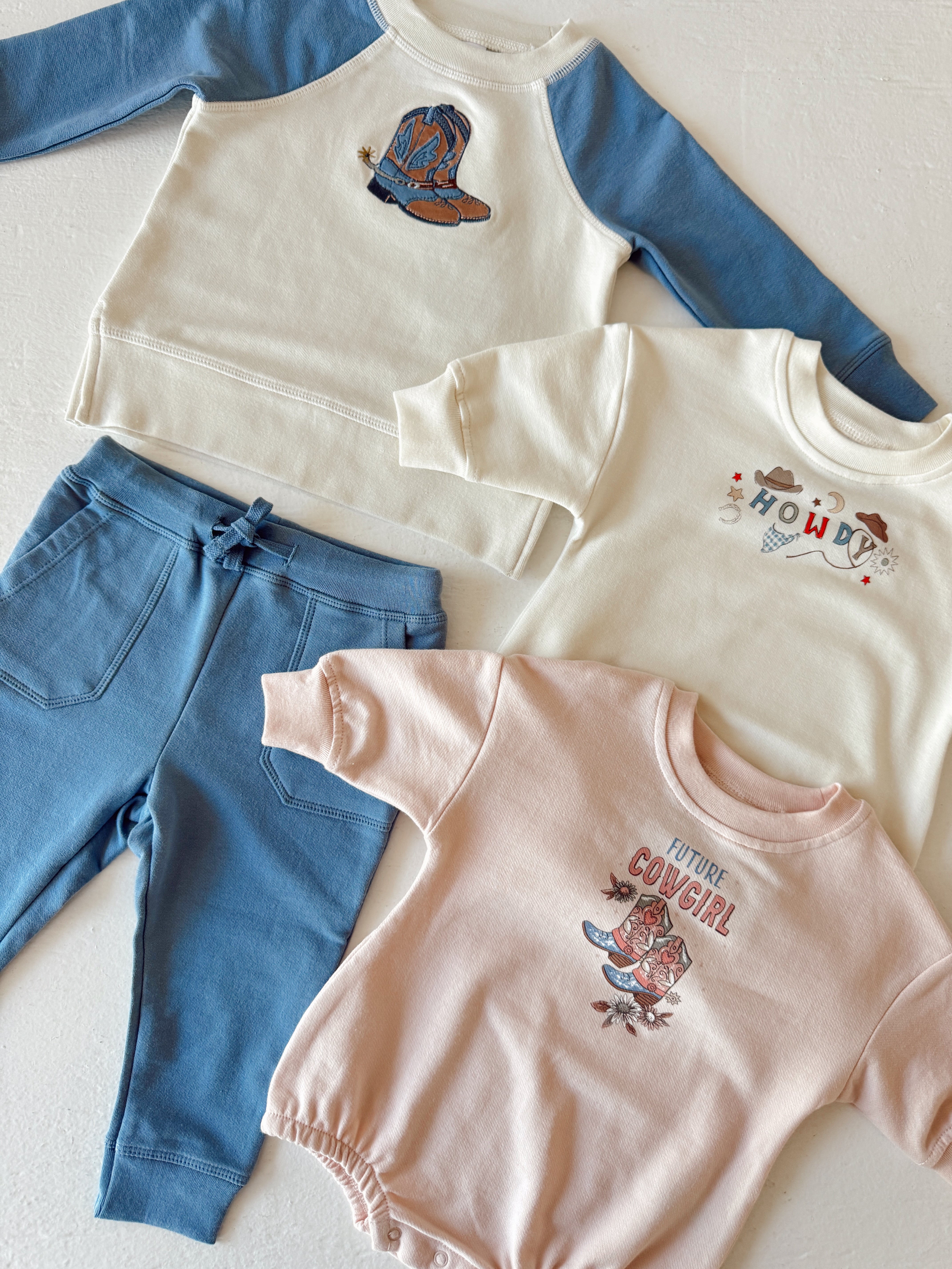 Kids' western-themed apparel: two sweatshirts, a onesie, and pants featuring cowboy motifs and playful designs.