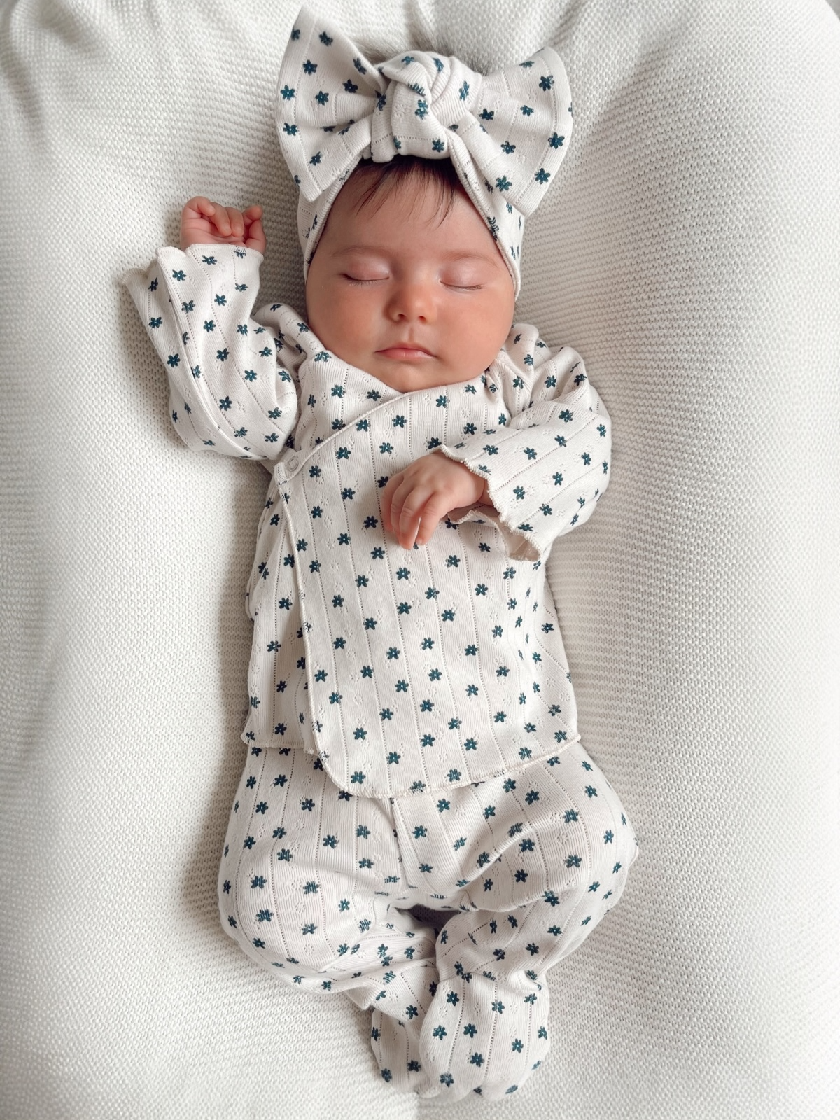 Sleeping baby in a soft outfit with floral patterns, wearing a matching headband, on a textured white surface.