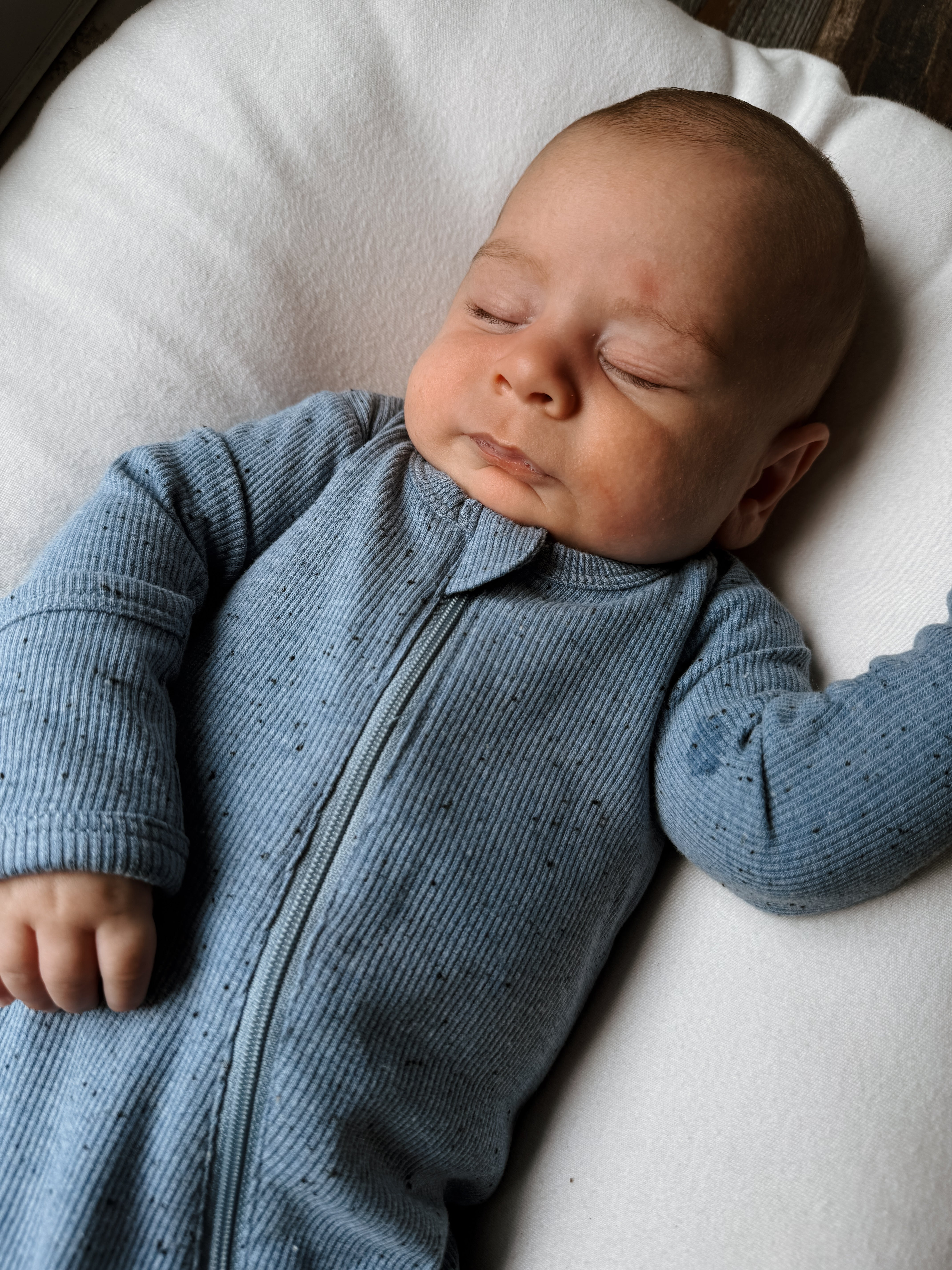 Sleeping baby in a blue ribbed outfit resting on a soft white surface.