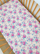 Floral crib sheet with pink roses and blue accents on a light background, displayed in a wooden crib.
