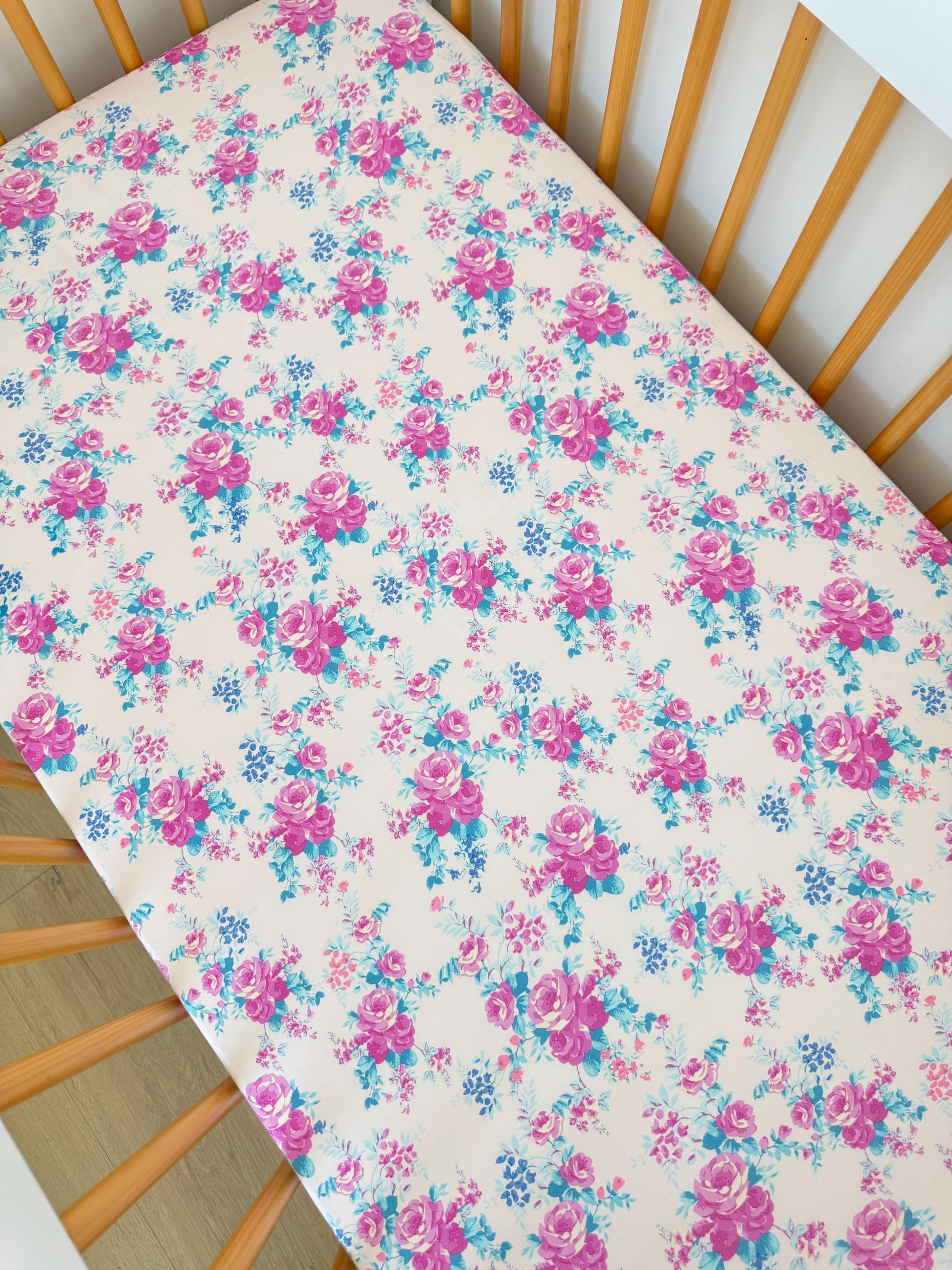 Floral crib sheet with pink roses and blue accents on a light background, displayed in a wooden crib.
