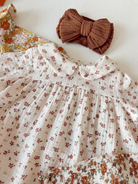 Floral baby dress with a collar and a brown fabric bow hair accessory on a light surface.
