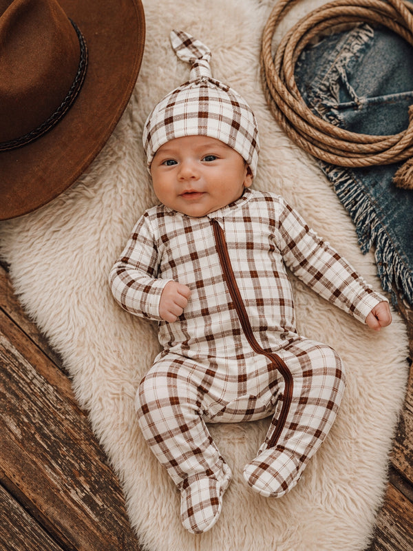 Smiling baby in a checkered outfit and hat, resting on a soft blanket surrounded by rustic decor.