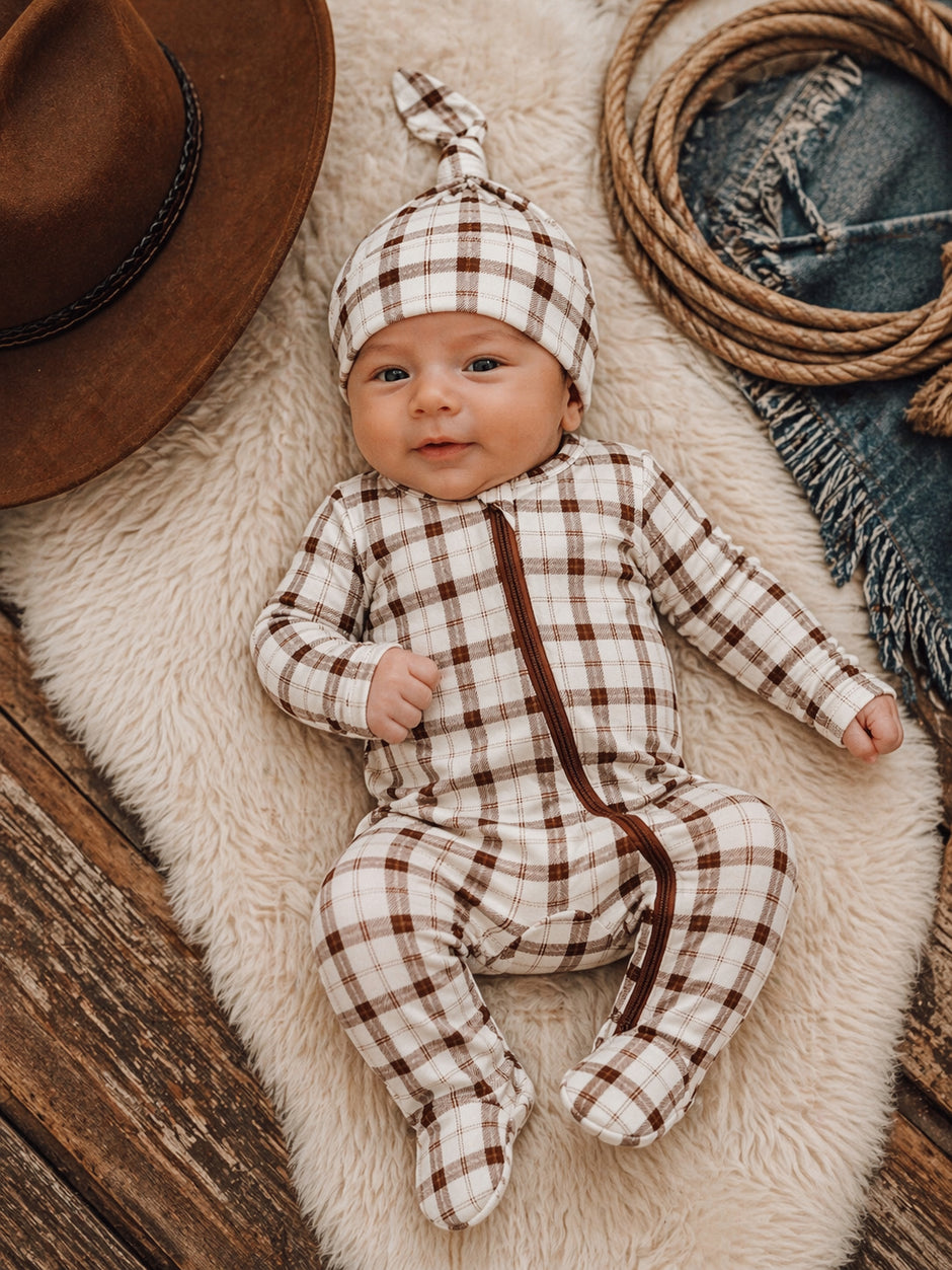 Smiling baby in a checkered outfit and hat, resting on a soft blanket surrounded by rustic decor.