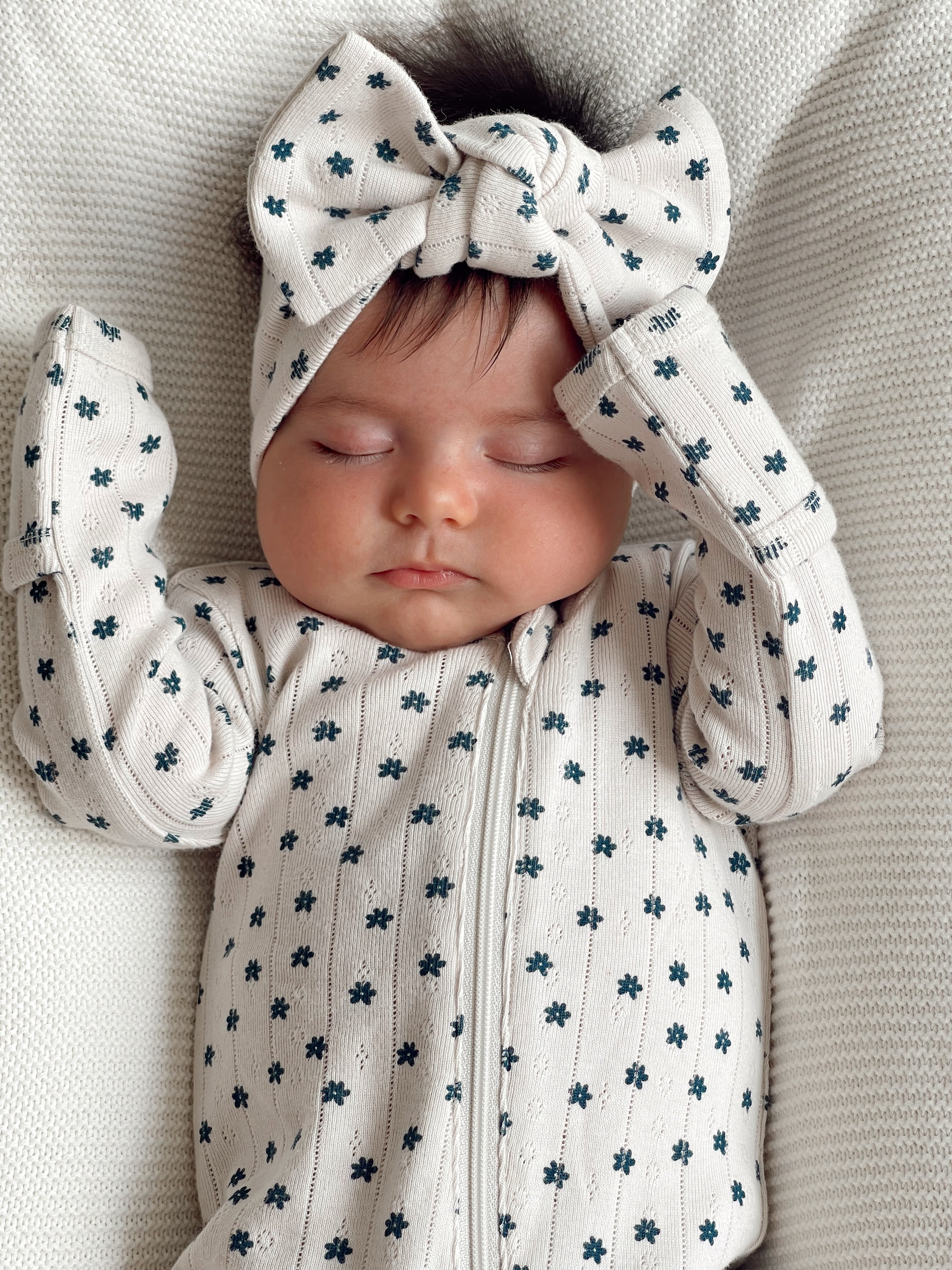 Baby asleep in floral print onesie and headband, resting on a soft textured blanket.