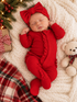Sleeping baby in red outfit with bow, surrounded by holiday decorations and a teddy bear on cozy blanket.