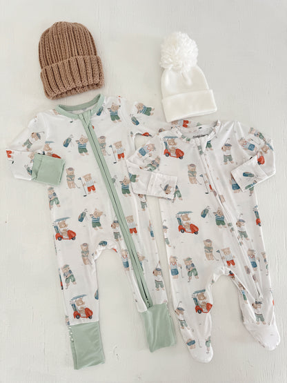 Two baby pajamas with bear patterns, paired with a brown knit hat and a white pom-pom beanie, on a light background.