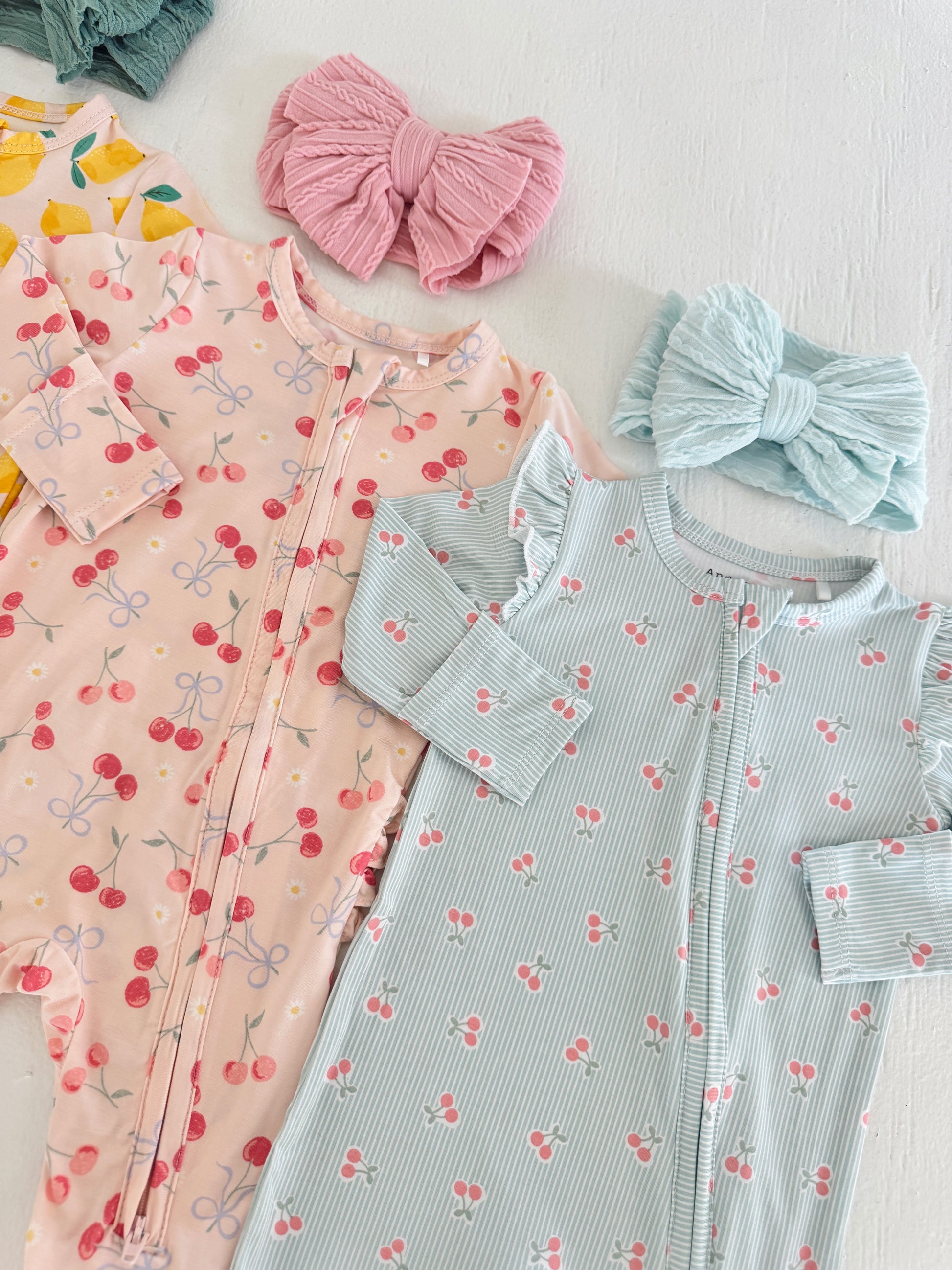 Two baby bodysuits with cherry patterns and coordinating hair bows in pink and blue on a white background.
