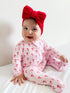 Smiling baby in a pink candy-cane outfit with a large red bow, sitting on a white bed.
