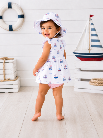 Smiling toddler in a sailboat-patterned dress and hat, standing near nautical decor in a bright, airy setting.