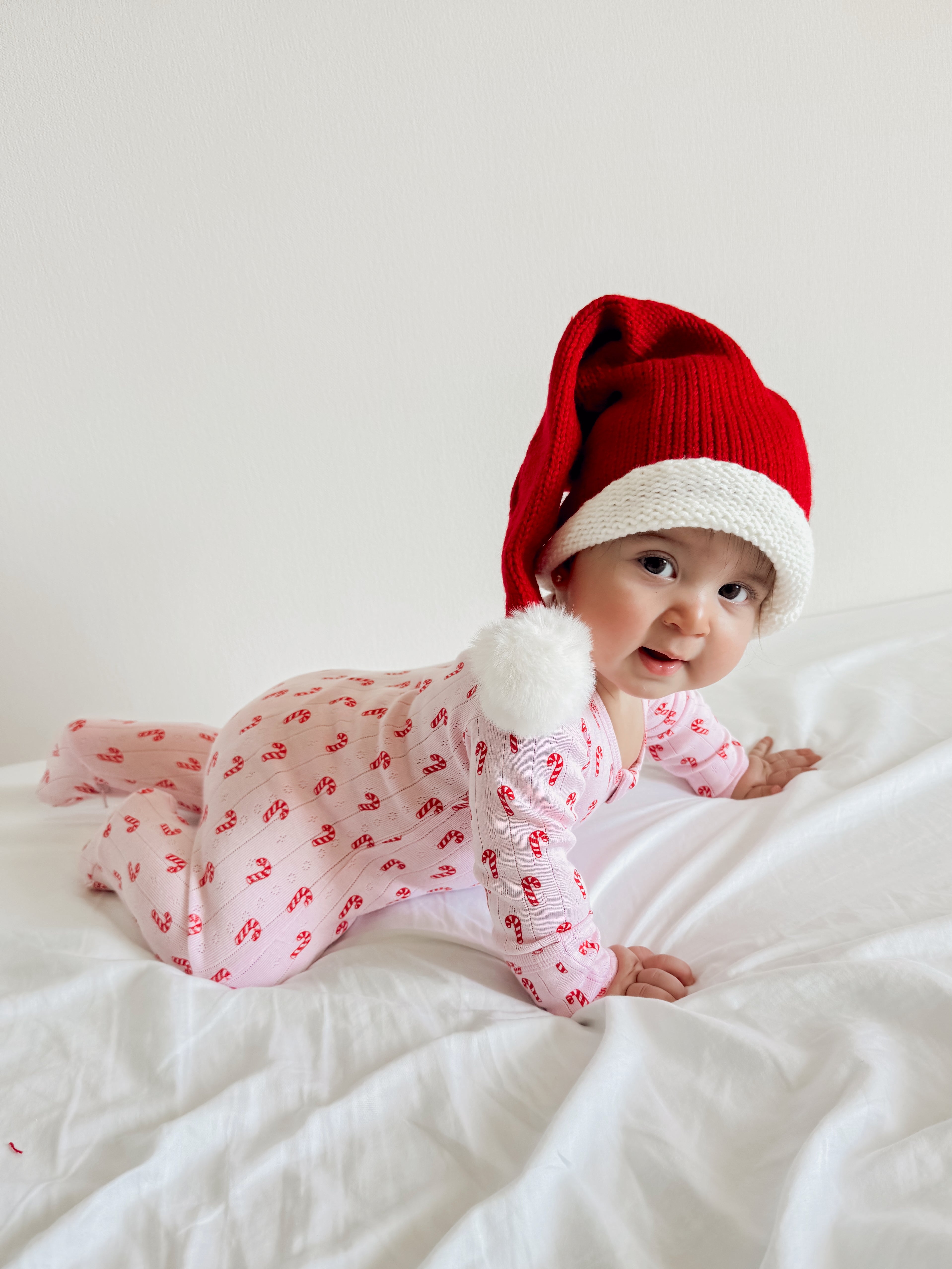 Infant in a red and white Santa hat crawling on a white bedspread, smiling brightly.