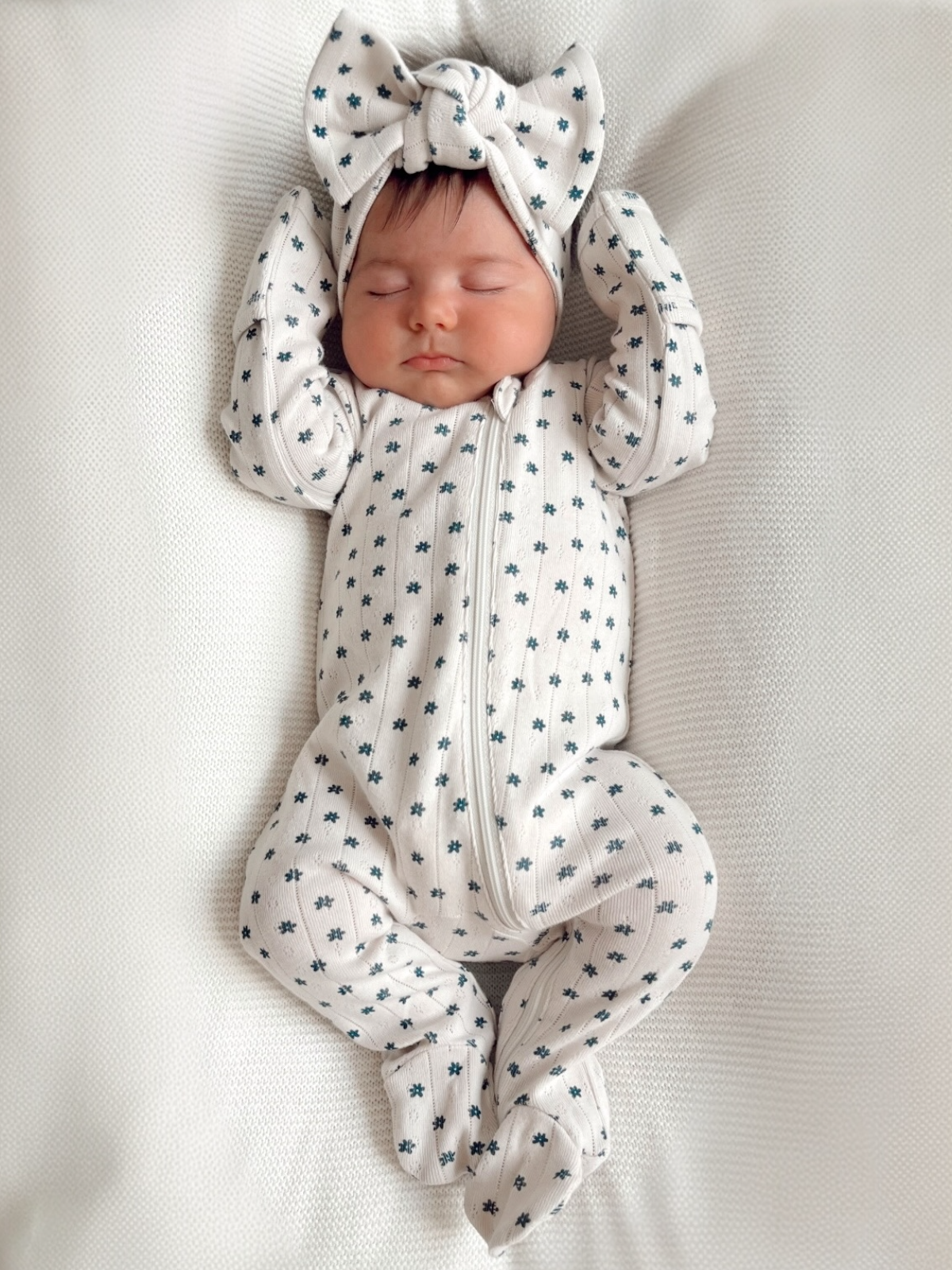 Sleeping baby in a floral-patterned onesie and headband, lying on a textured white surface.