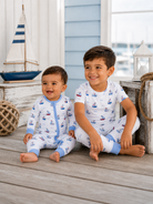 Two children in matching sailboat pajamas sit on a wooden floor, smiling, with nautical decor in the background.
