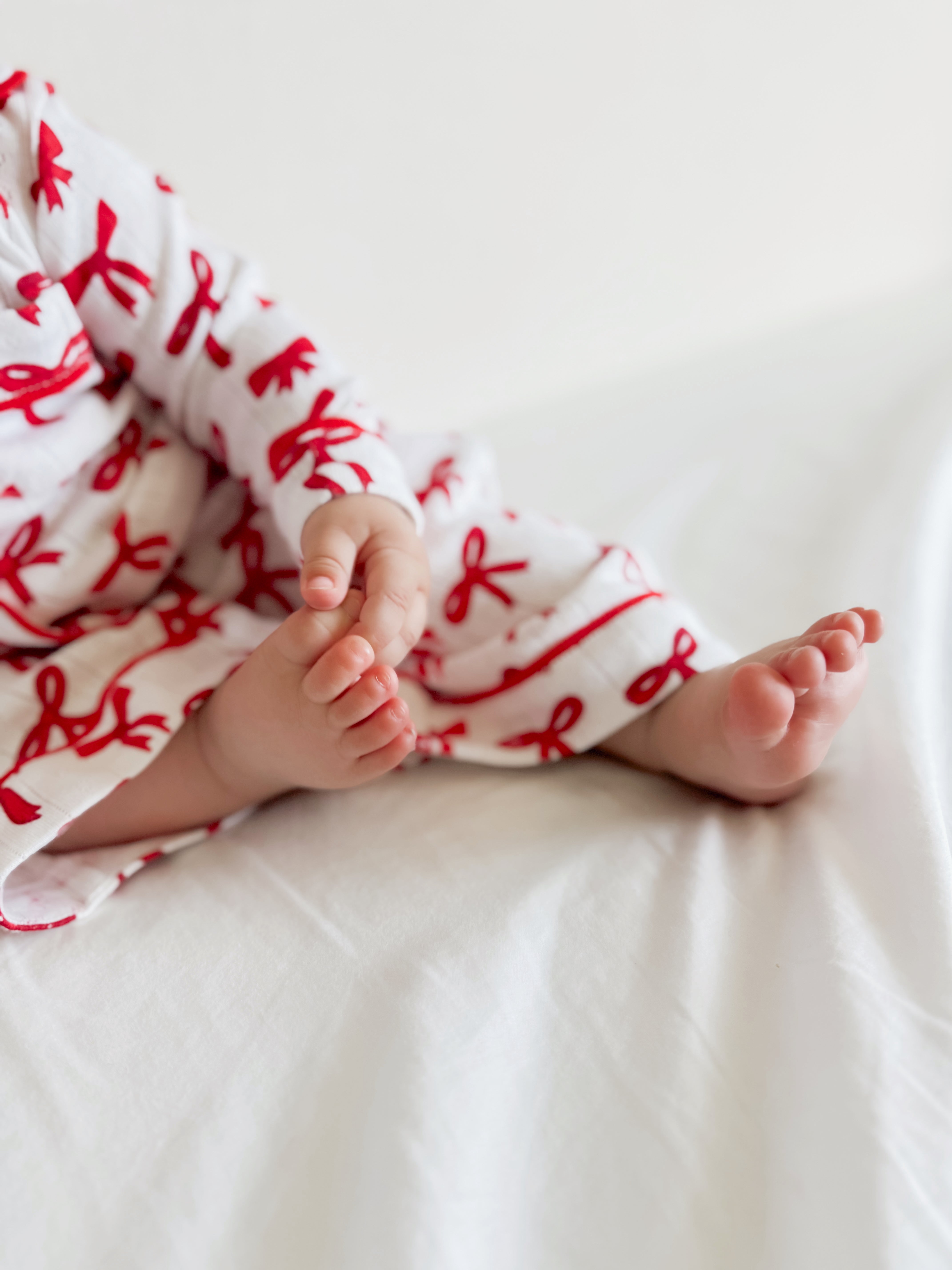 Baby feet and hands playfully resting on a white bed, wearing a festive red and white outfit with bows.