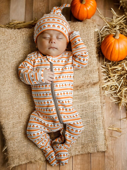 Sleeping baby in pumpkin-patterned outfit, surrounded by pumpkins on a straw mat. Warm autumn theme.