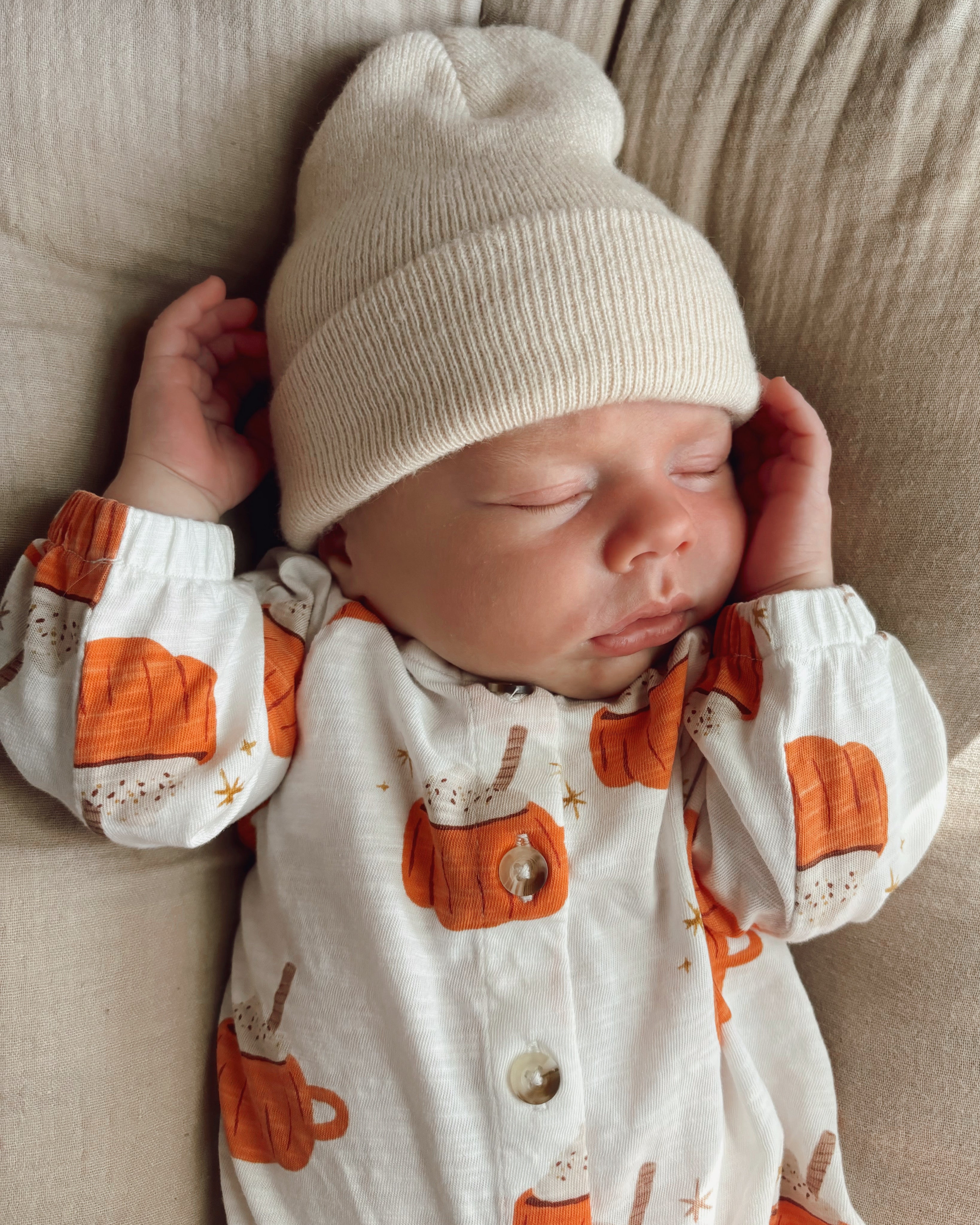 Sleeping baby in a pumpkin-patterned outfit and cream beanie, resting peacefully on a beige surface.