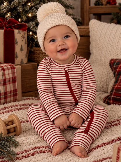 Smiling baby in striped pajamas and a knit hat, sitting on a cozy rug among holiday decorations.