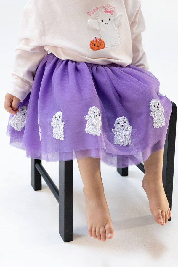 Child wearing a purple tulle skirt with ghost appliqués, seated on a black chair, feet barefoot.
