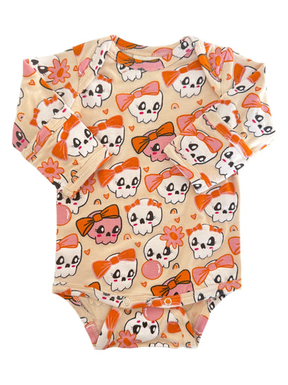 Long-sleeve baby onesie with a playful skull and bow pattern in pastel colors against a cream background.