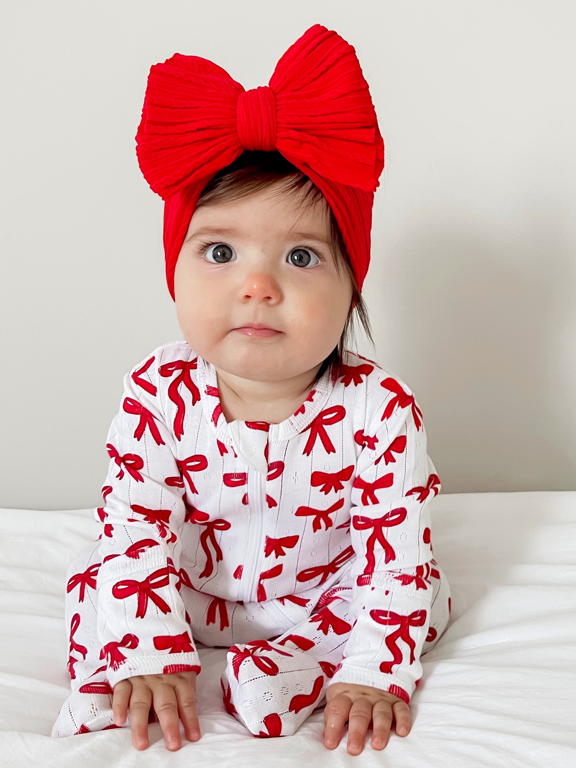 Baby girl in red bow headband and festive outfit with bows, sitting on a white bedspread, looking curiously at the camera.