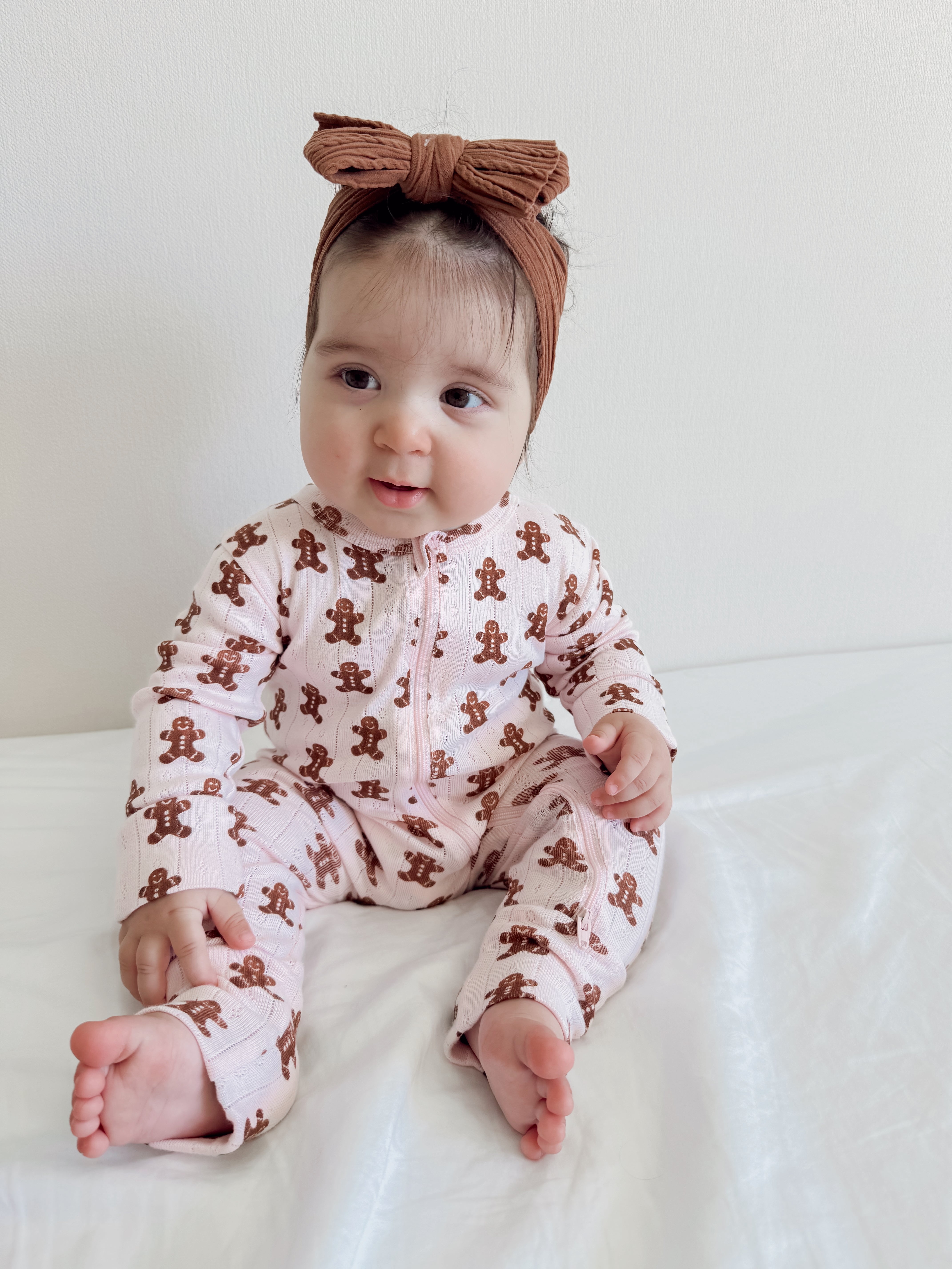 Baby in gingerbread-print pajamas and a brown bow headband, sitting on a white blanket, smiling.