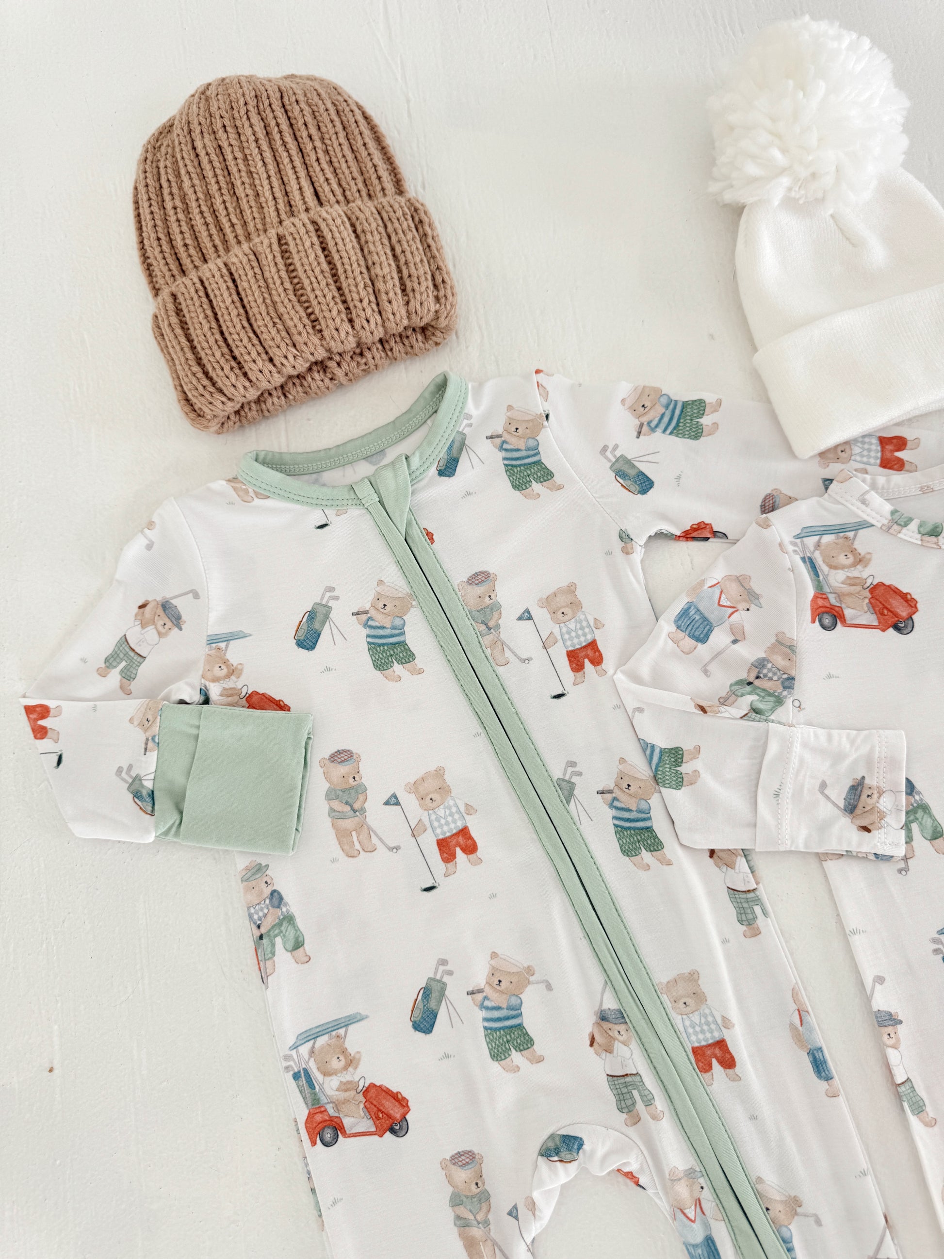 Baby pajamas with bear print, green accents, and two knit hats, one white with a pom-pom and one brown.