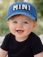 Smiling baby wearing a blue cap with "MINI" on it, sitting outdoors, with soft green background.
