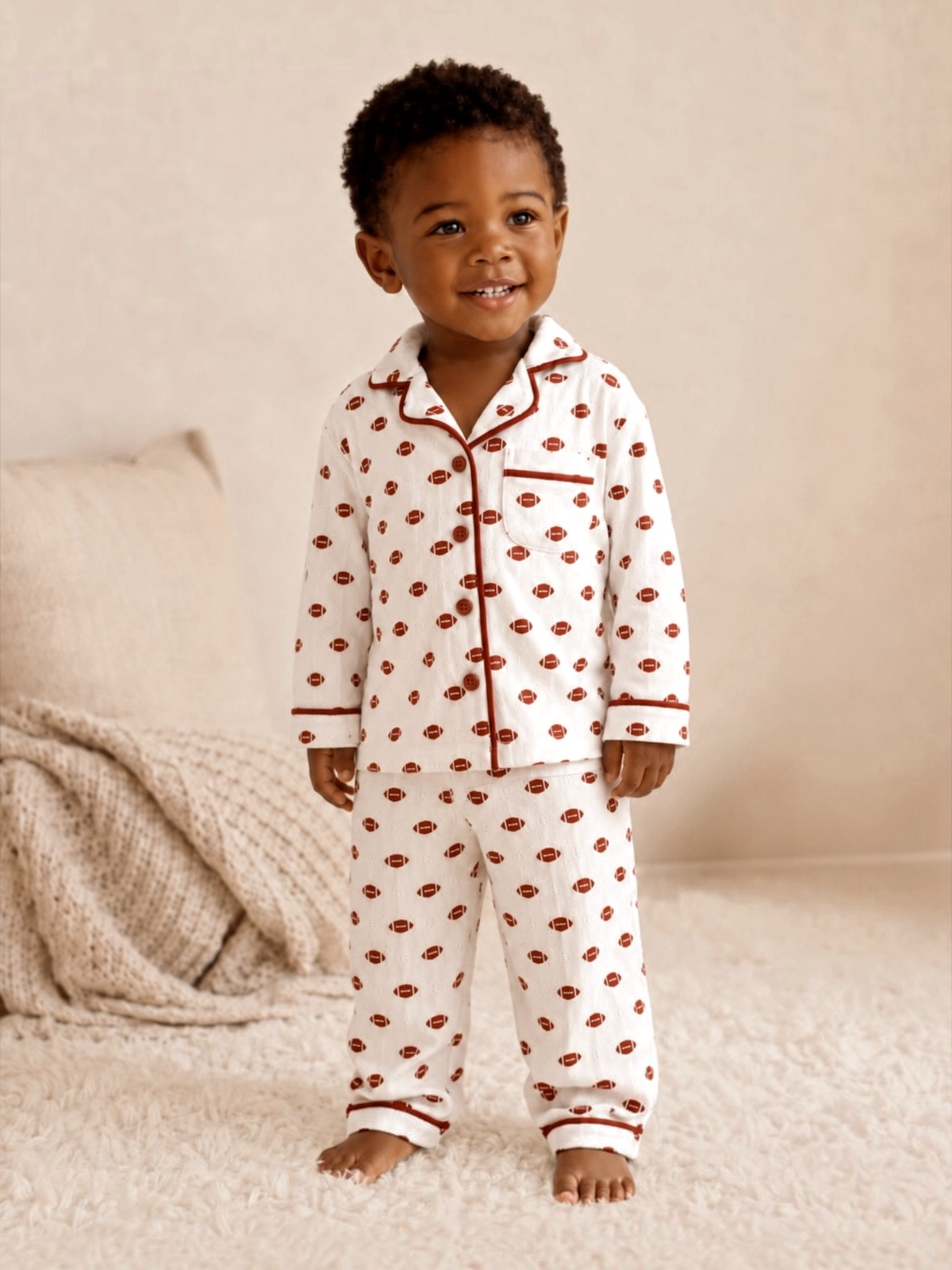 Smiling young boy in white pajamas with red patterns, standing on a cozy rug in a softly lit room.