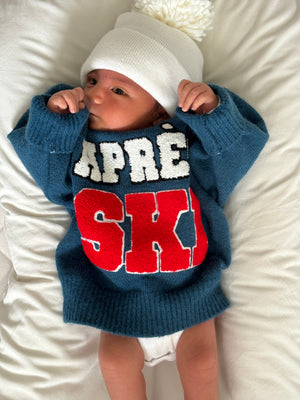 Baby wearing a blue sweater with "APRES SKI" text and a white beanie, lying on a soft white surface.