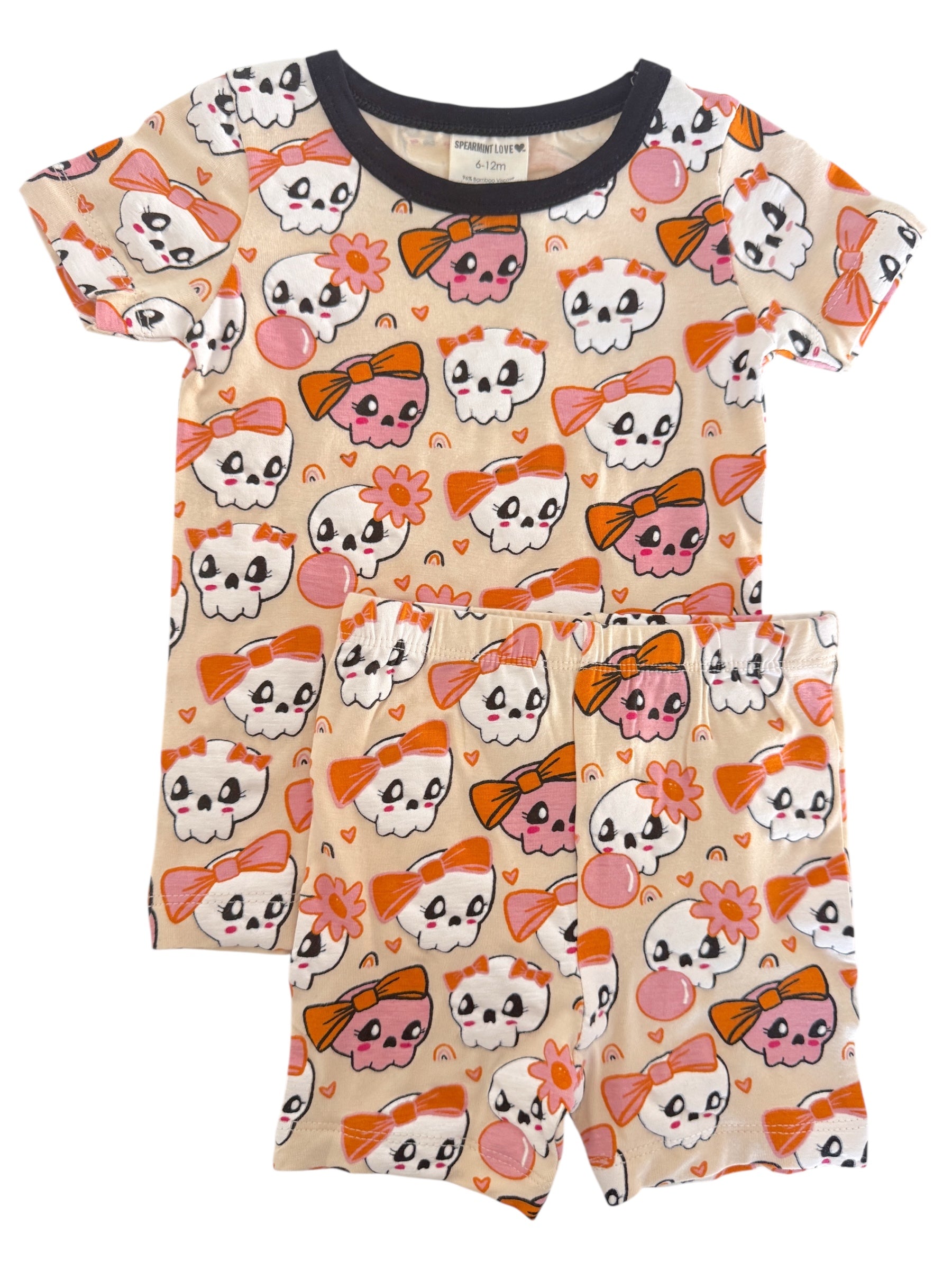 Kid's short pajama set featuring cute skulls with bows and flowers in playful colors against a beige background.