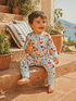 Smiling child in colorful play suit, sitting on stone steps with potted plants and a serene outdoor background.