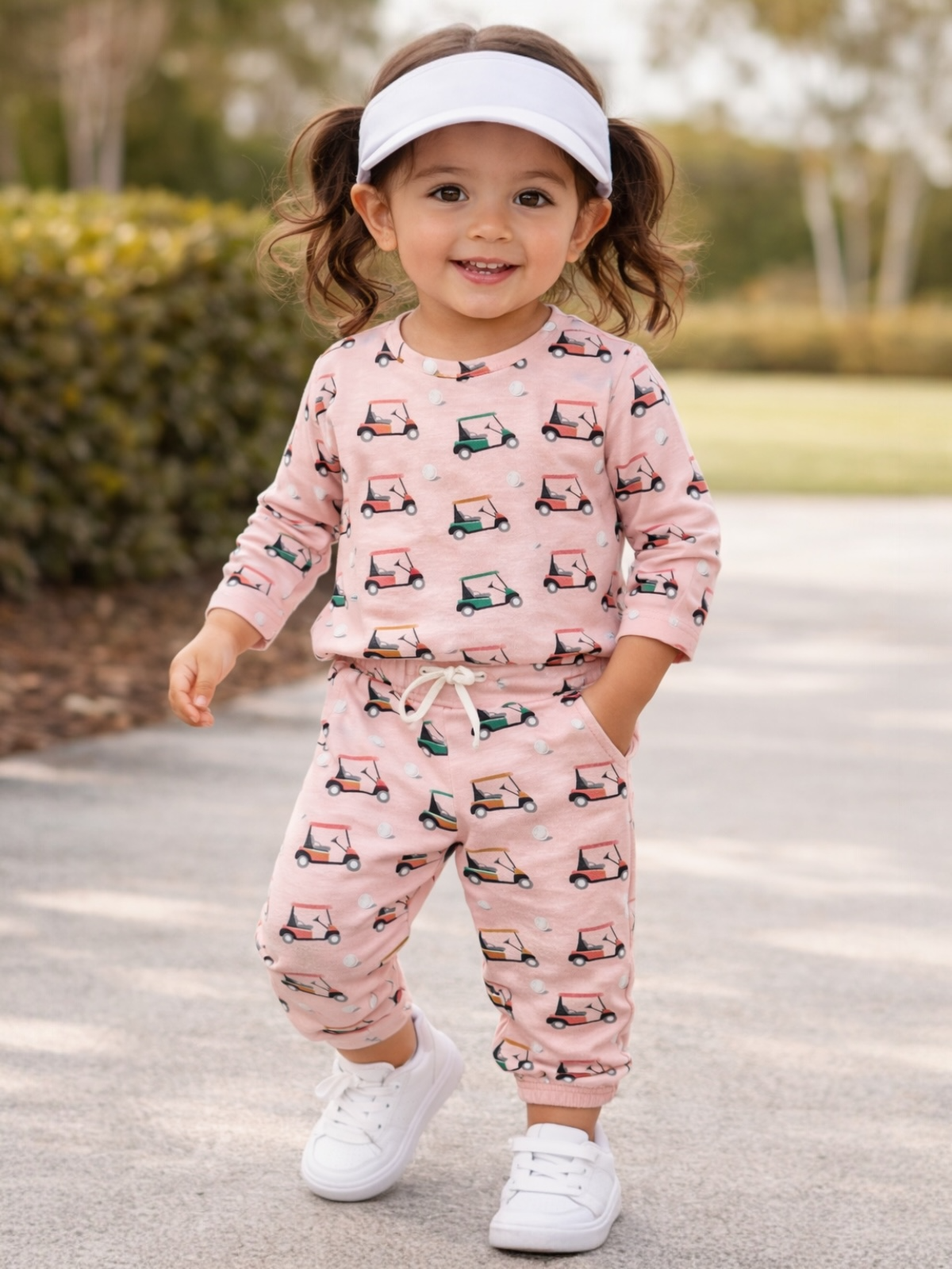 Smiling toddler in pink golf cart-patterned outfit, walking on a path, wearing a white visor and sneakers.