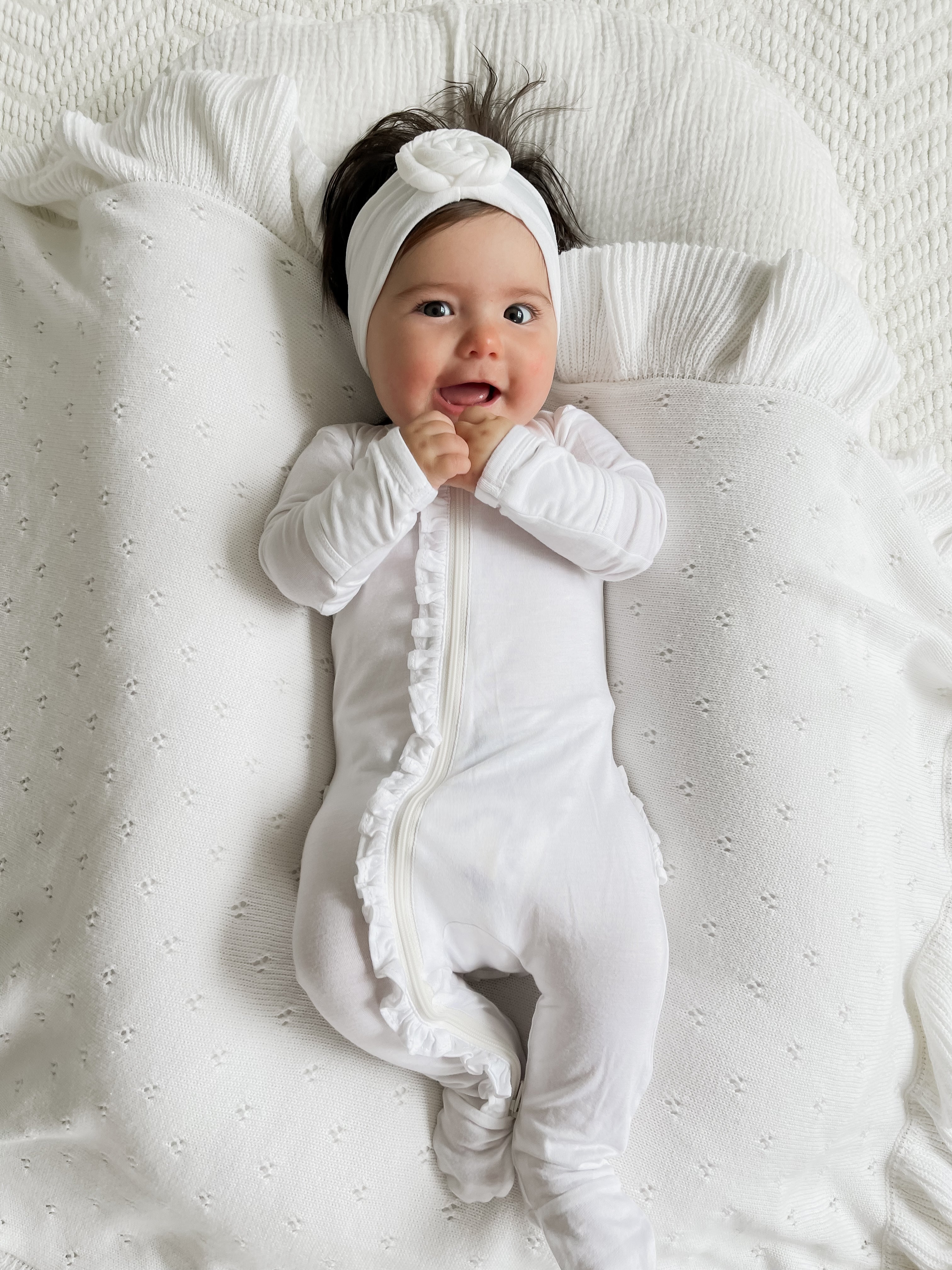 Smiling baby in a white outfit and headband, lying on a textured white blanket with ruffles.