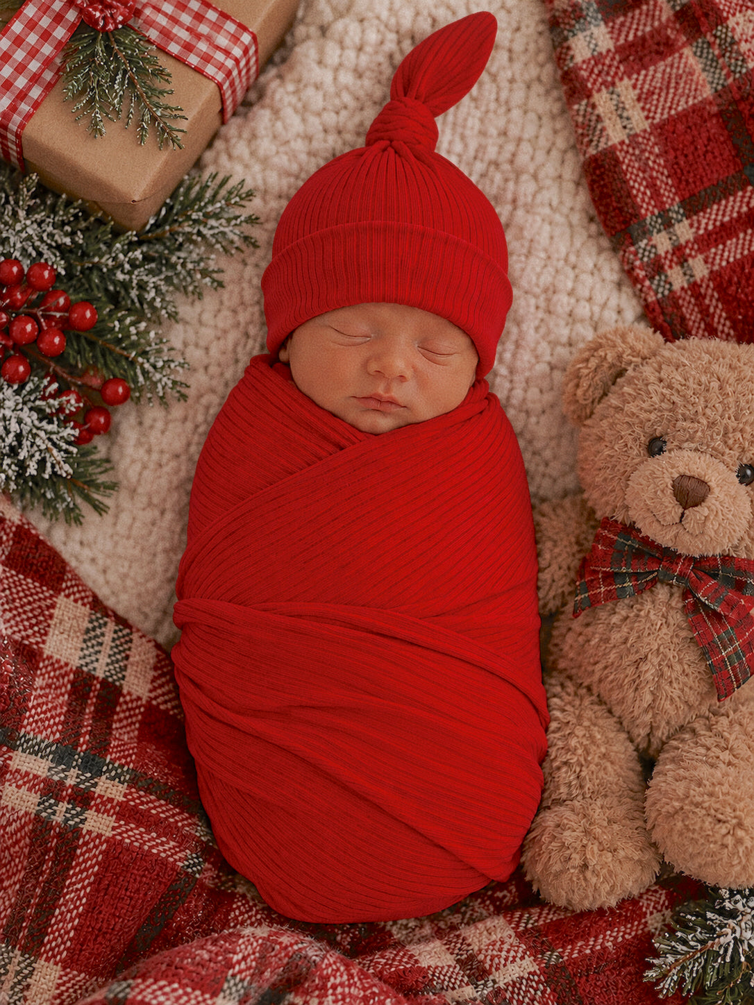 Newborn swaddled in red, sleeping peacefully beside a teddy bear on a cozy plaid blanket and festive decor.