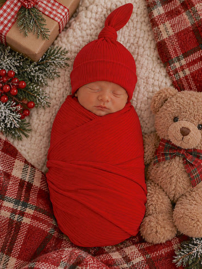 Newborn swaddled in red, sleeping peacefully beside a teddy bear on a cozy plaid blanket and festive decor.