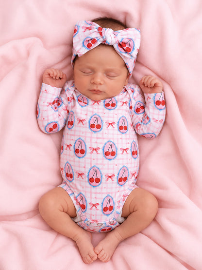 Smiling baby in cherry-print onesie and bow, resting on a pink blanket.