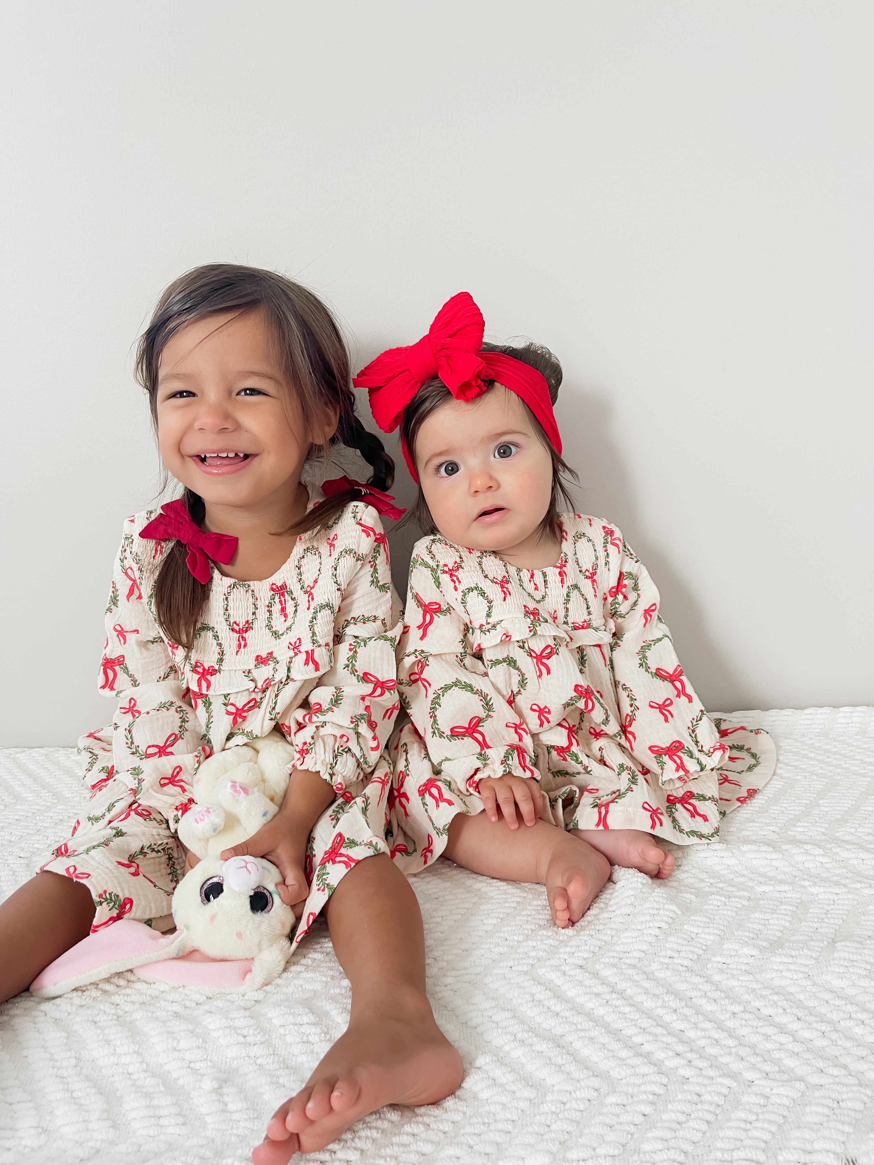 Two young girls in matching holiday dresses sit on a white blanket, one holding a stuffed bunny toy.