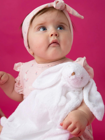 Baby in a pink outfit holding a white bunny blanket, set against a bright pink background.