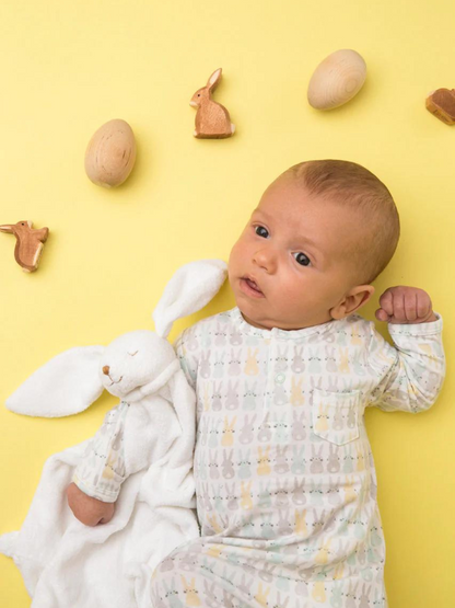 Baby dressed in rabbit-patterned pajamas holds a soft bunny toy on a yellow background with wooden decorations.