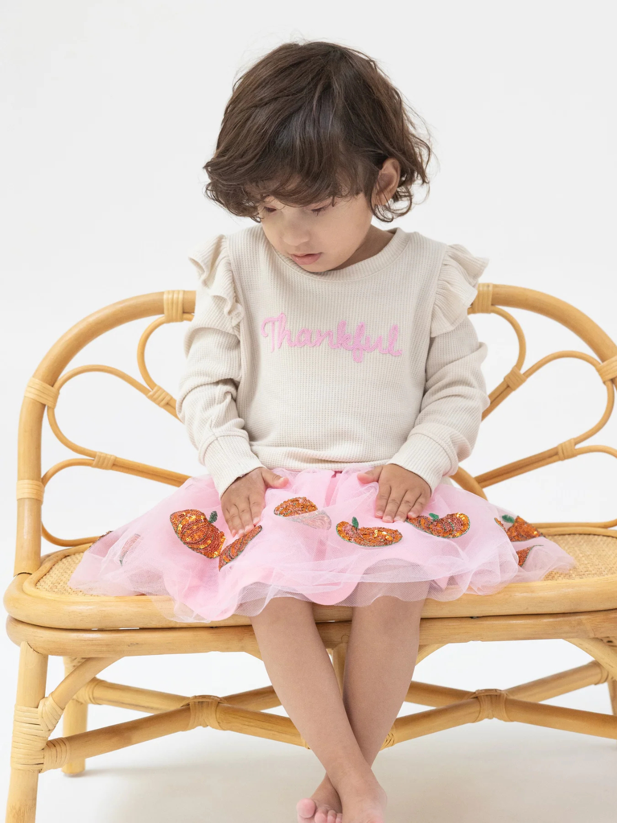 Child sitting on a rattan chair, wearing a "Thankful" sweater and a pink tutu skirt with colorful decorations.