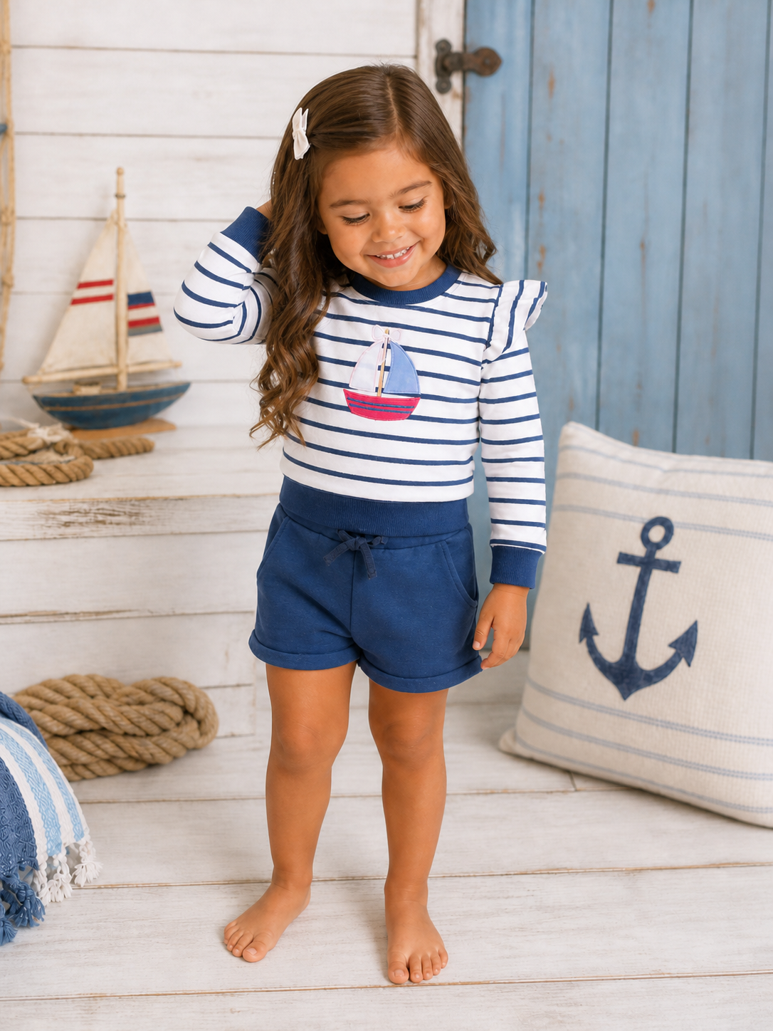 Smiling girl in a striped outfit with a boat graphic, standing near nautical decor in a bright, cozy room.