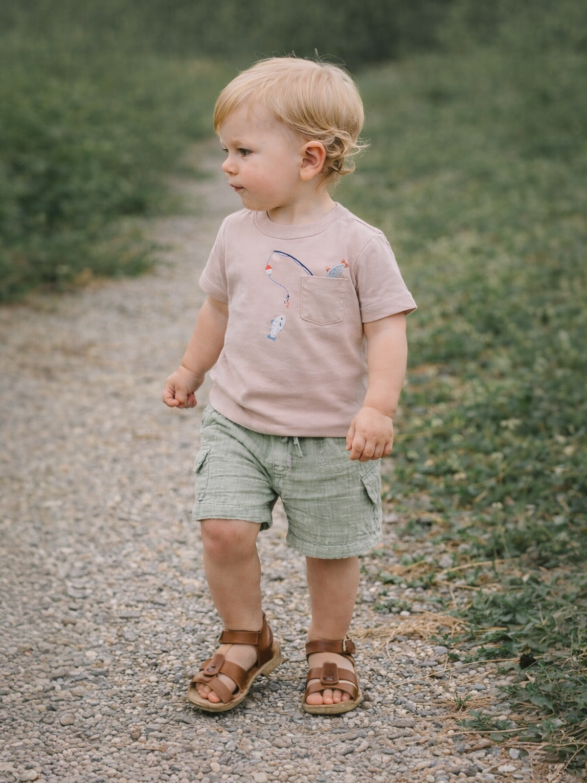Toddler in casual wear walking on a gravel path surrounded by greenery.