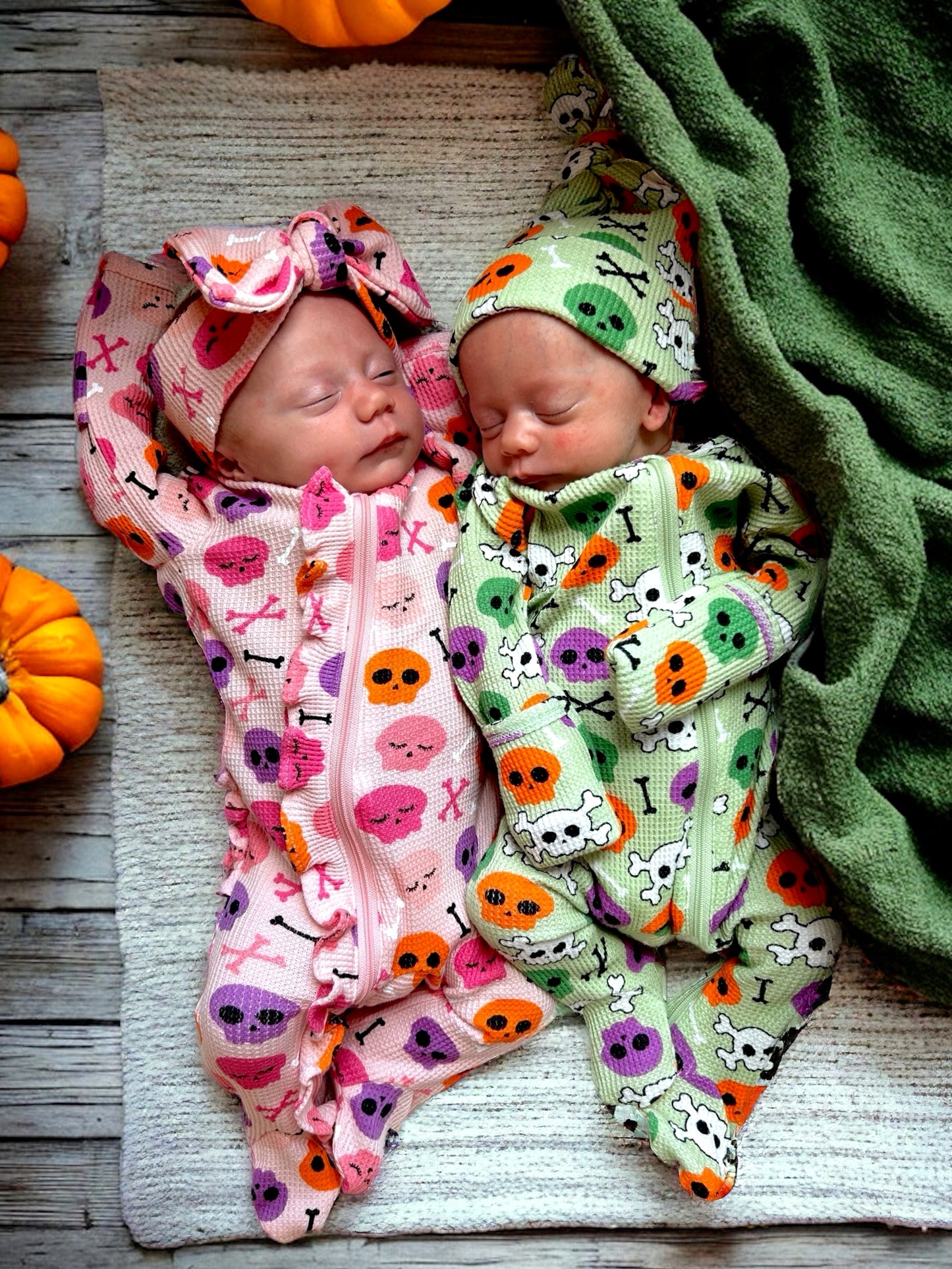Two newborns in Halloween-themed outfits sleep on a textured blanket, surrounded by pumpkins and a green blanket.
